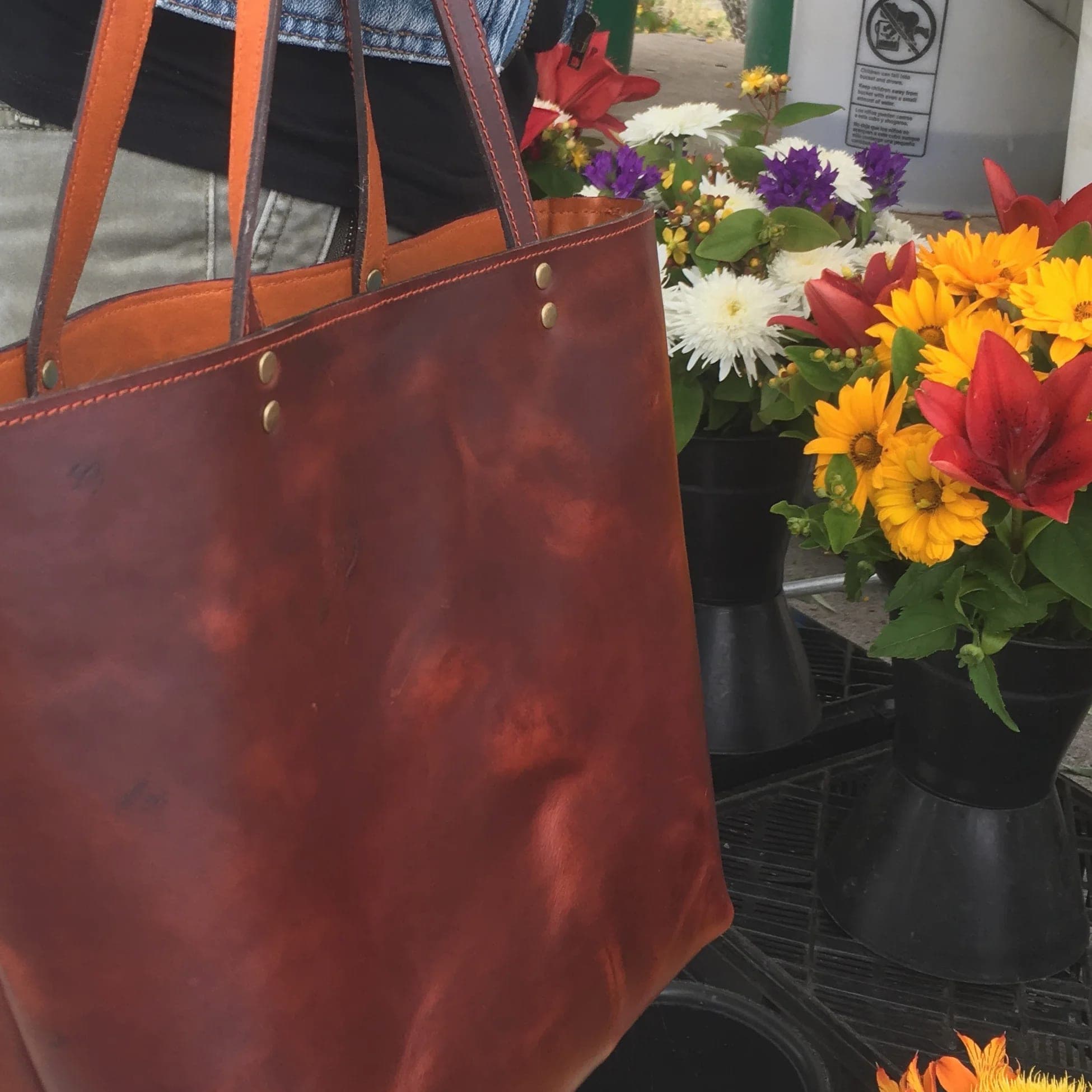 A reddish-brown leather tote with tan straps and brass rivets, at a flower stall beside buckets of dahlias
