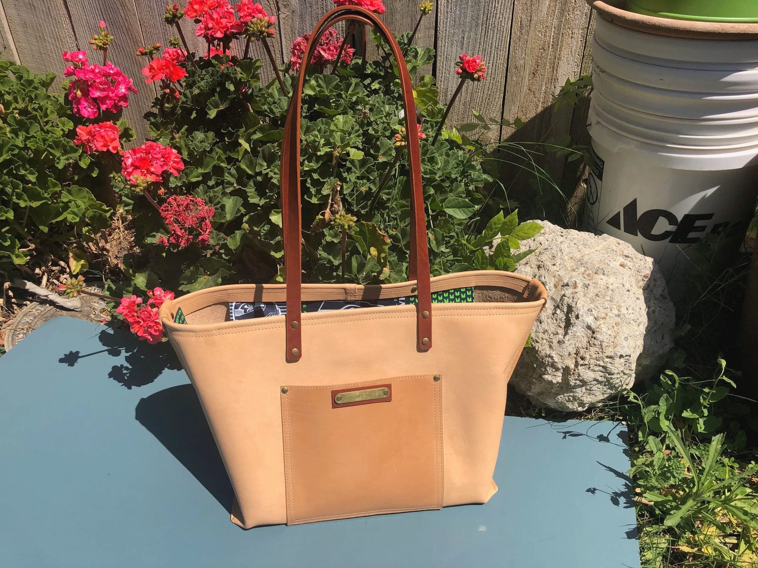 Natural tan leather tote with a stamped nameplate, on a stone ledge among red geraniums