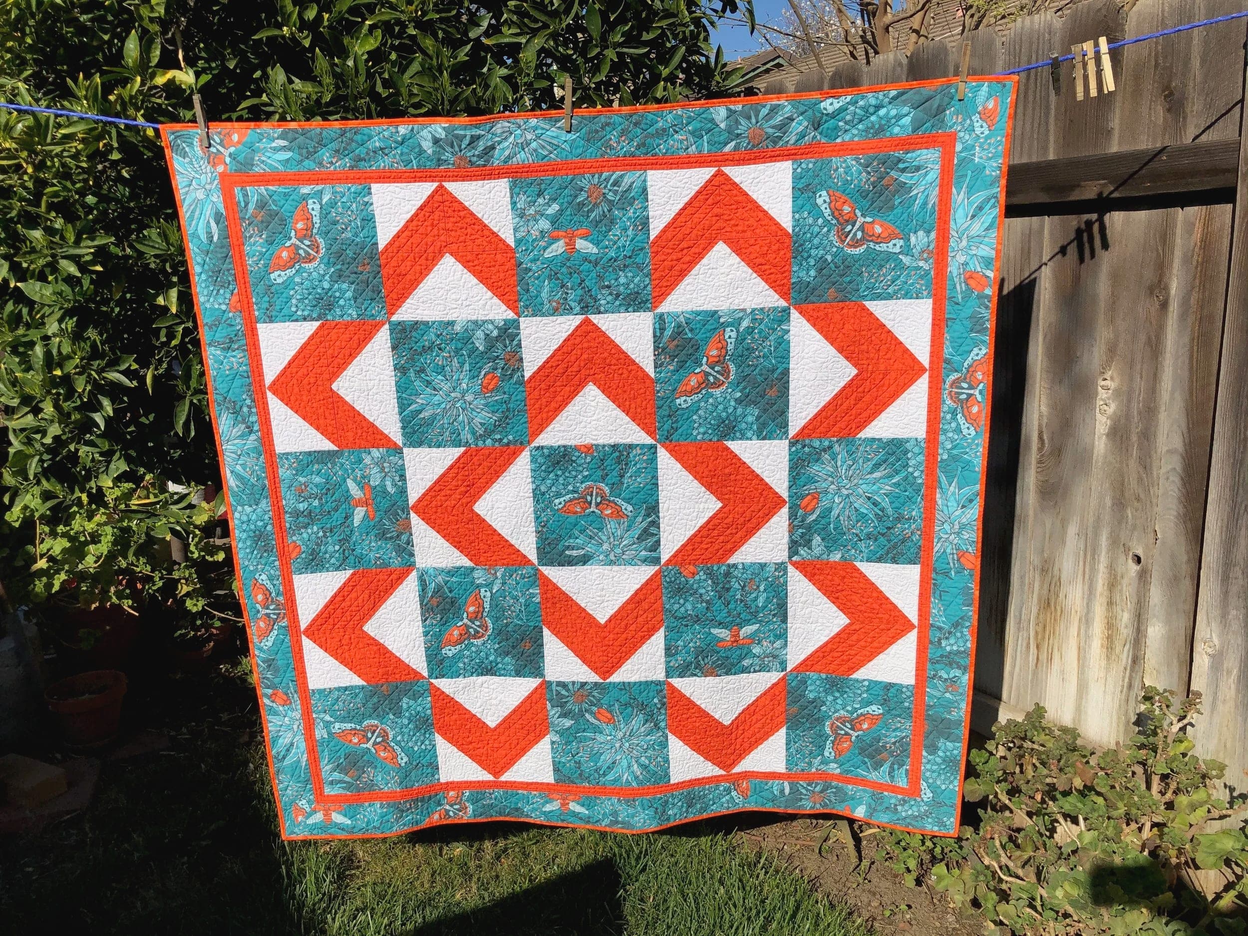 Patchwork quilt in teal, orange and white with a repeating arrow pattern, hanging on a garden line