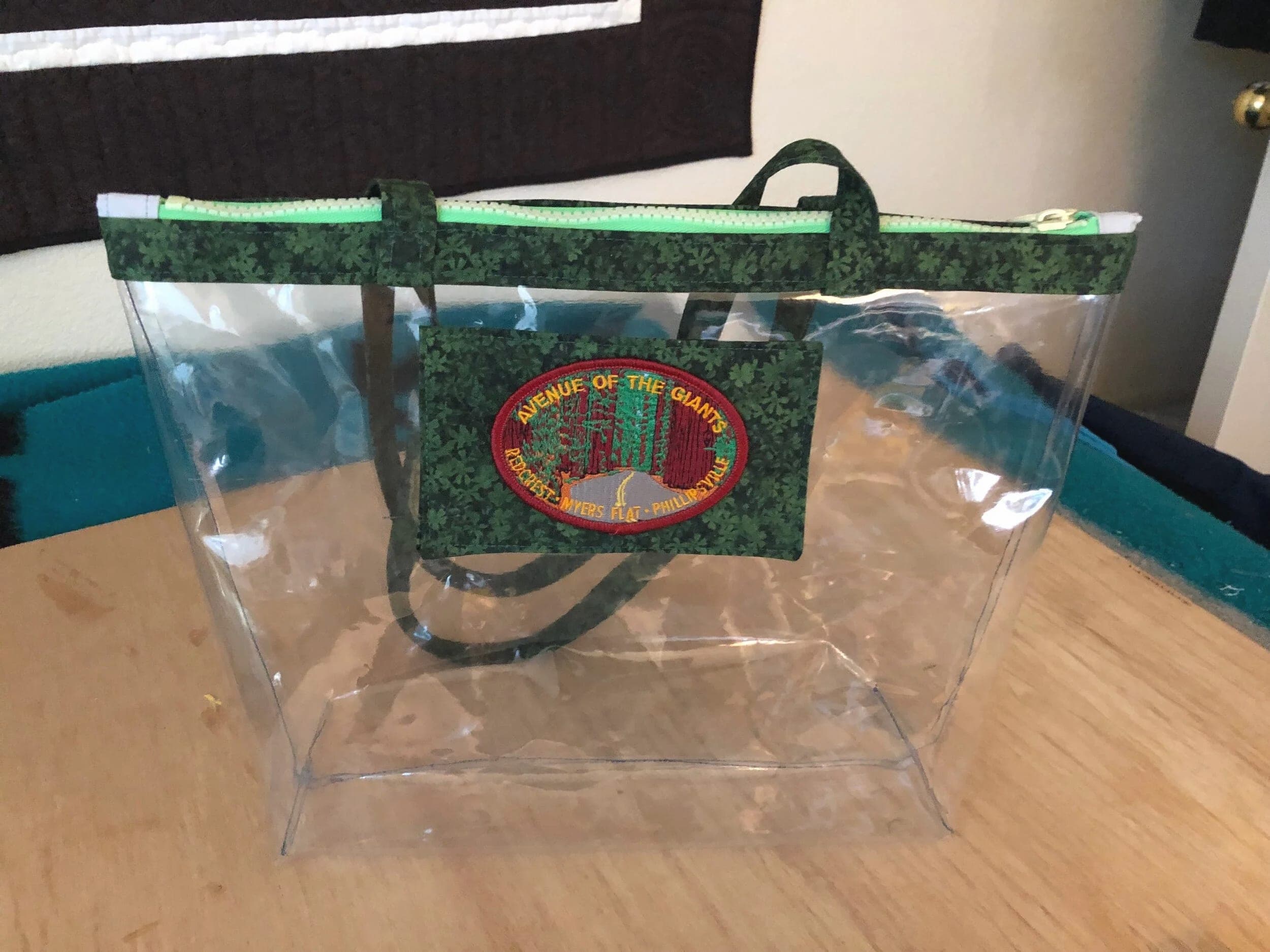 Clear vinyl tote with green print trim and handles and an embroidered patch, on a wood floor