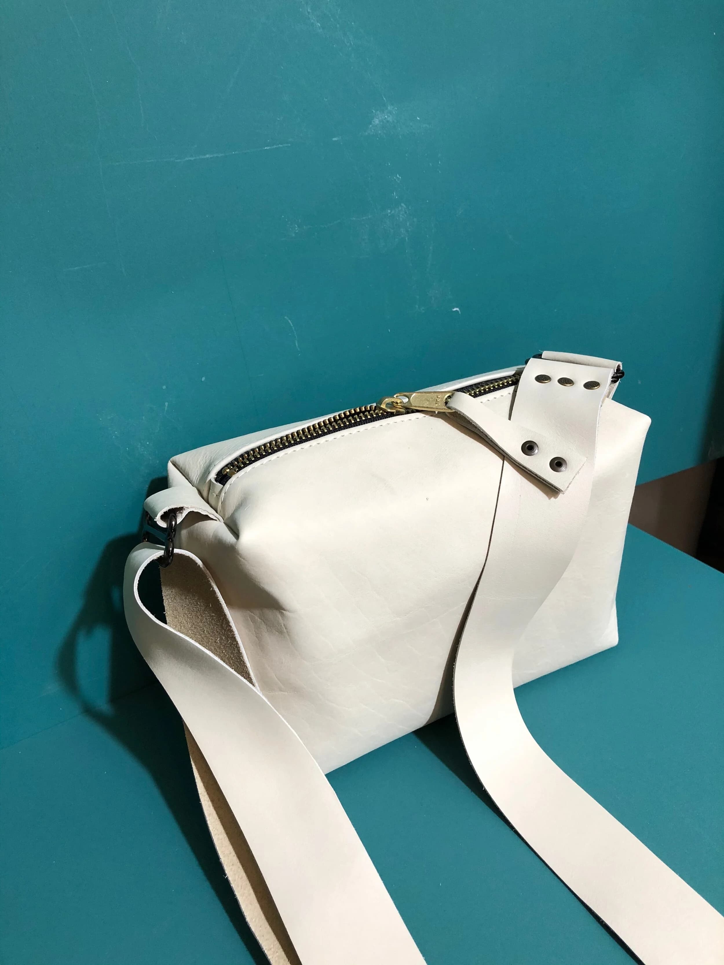 White leather zip bag with a long strap and silver hardware, on a teal surface