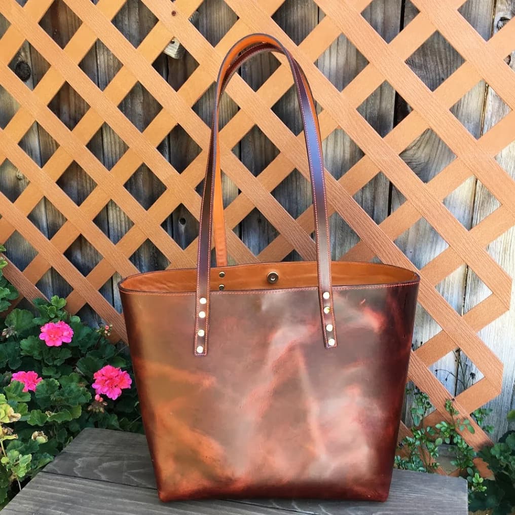 Reddish-brown leather tote with tan straps and brass rivets, on a bench against a lattice fence