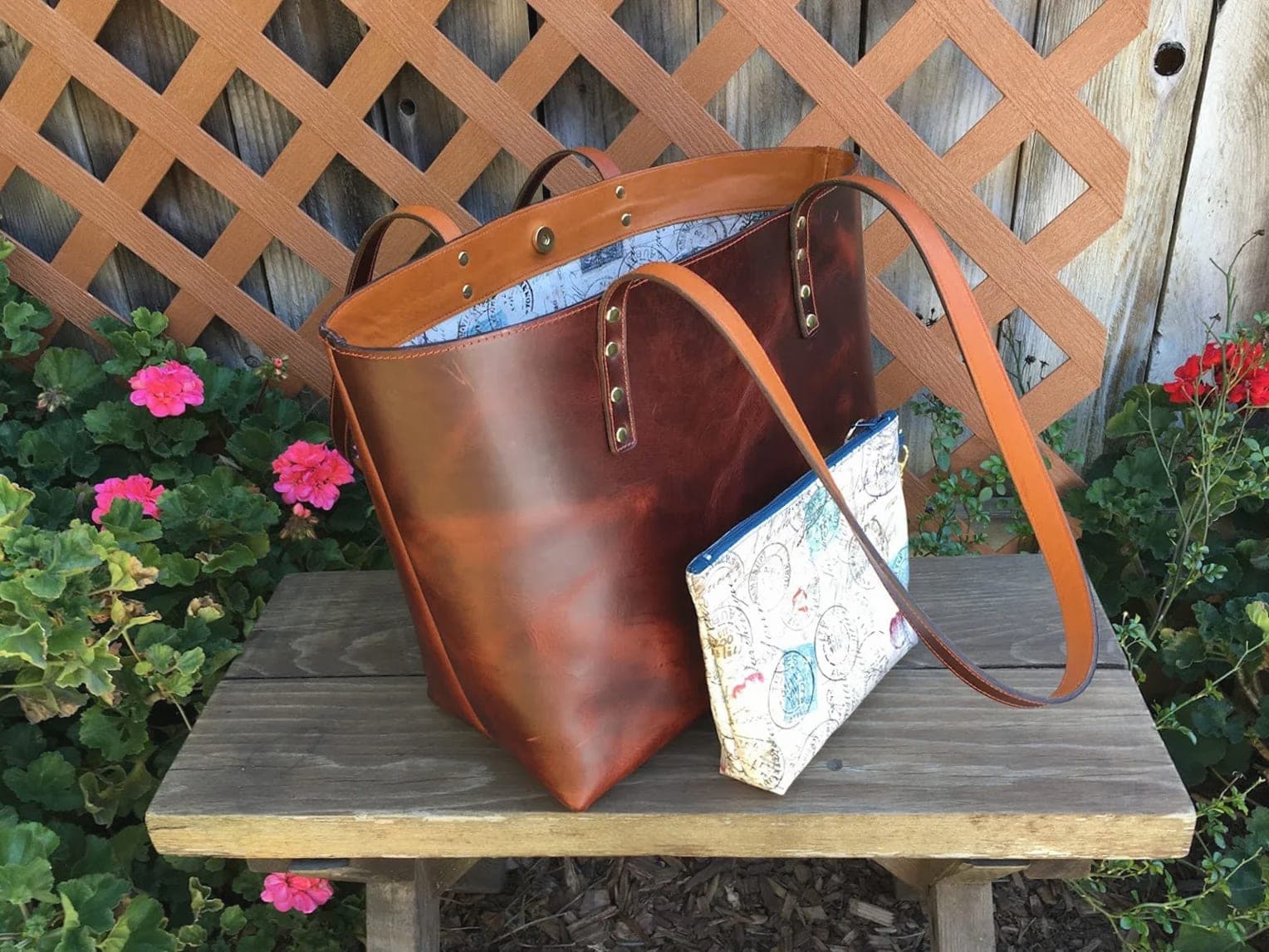 A reddish-brown leather tote on a garden bench beside a matching map-print zip pouch
