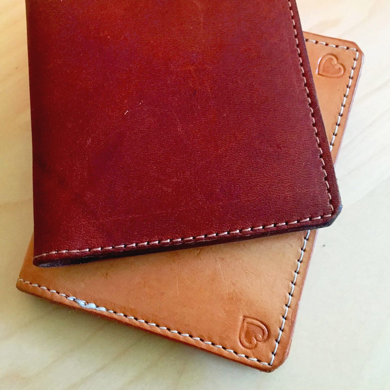 Two handmade leather bifold wallets, one oxblood and one natural tan, with white contrast stitching