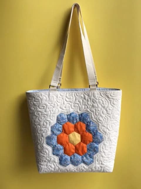 Quilted white tote with a blue and orange hexagon flower motif, hanging against a yellow wall